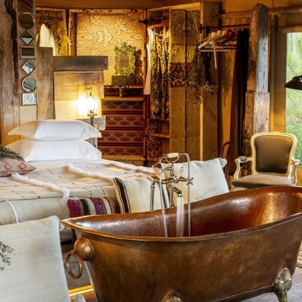 A cozy, stylish bedroom featuring a vintage copper bathtub as a centerpiece, surrounded by rustic decor, patterned textiles, and soft lighting. A neatly made bed with white linens is visible, along with a wardrobe displaying colorful garments.