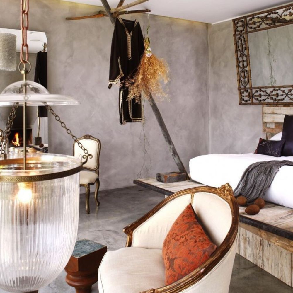 A stylish interior of a modern bedroom featuring a gray wall, a wooden bed platform, and a hanging mirror. A vintage armchair with an orange cushion sits nearby, alongside a unique pendant light. Decorative items include dried plants and a rustic tree branch.