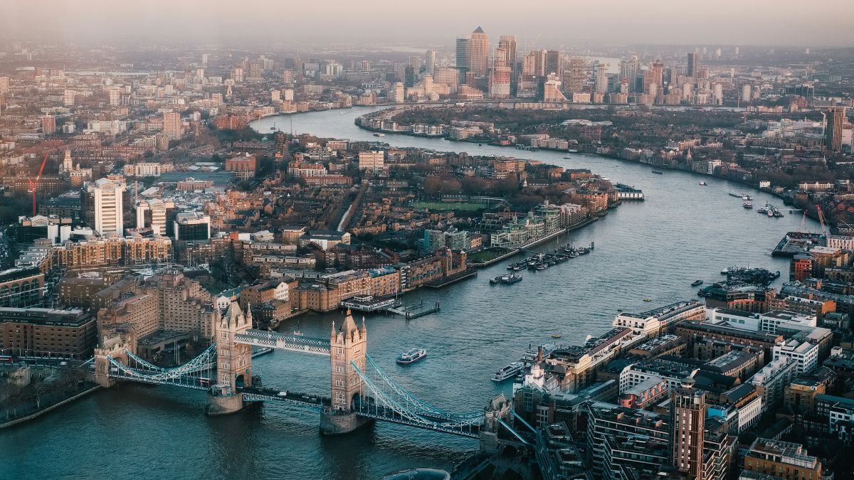 Aerial view of London showing the River Thames winding through the city. Tower Bridge stands prominently, while the skyline features modern skyscrapers in the background, with boats visible on the river.