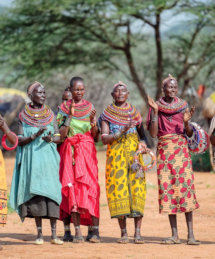 A group of five women in traditional attire, standing together outdoors. They are wearing brightly colored dresses and elaborate beaded jewelry, clapping and smiling, with a background of trees and a dusty ground.