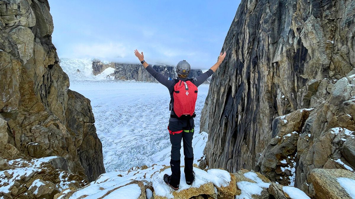 A person stands on a rocky ledge with arms raised, overlooking a vast glacier surrounded by towering cliffs. Snow covers the ground, and the sky is partly cloudy.