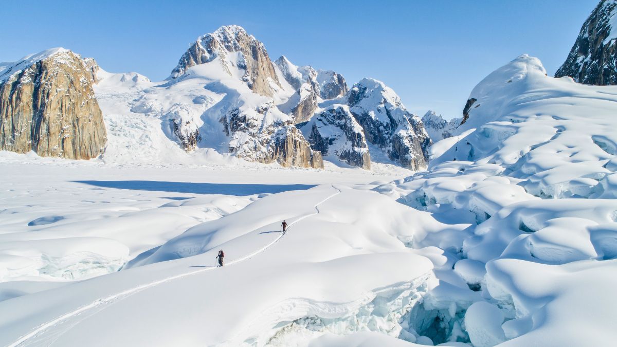 Two hikers trek across a vast expanse of snow and ice, surrounded by towering, rugged mountains and bright blue sky. The landscape features deep crevasses and glacial formations, creating a stunning winter scene.