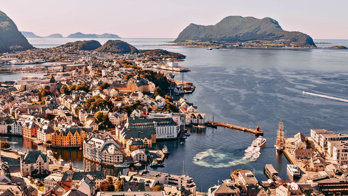 A panoramic view of a coastal town with colourful buildings lining the waterfront. The scene features a mix of residential and commercial structures, boats in the harbour, and distant mountains in the background. The water is calm, reflecting the surrounding landscape under clear blue skies.