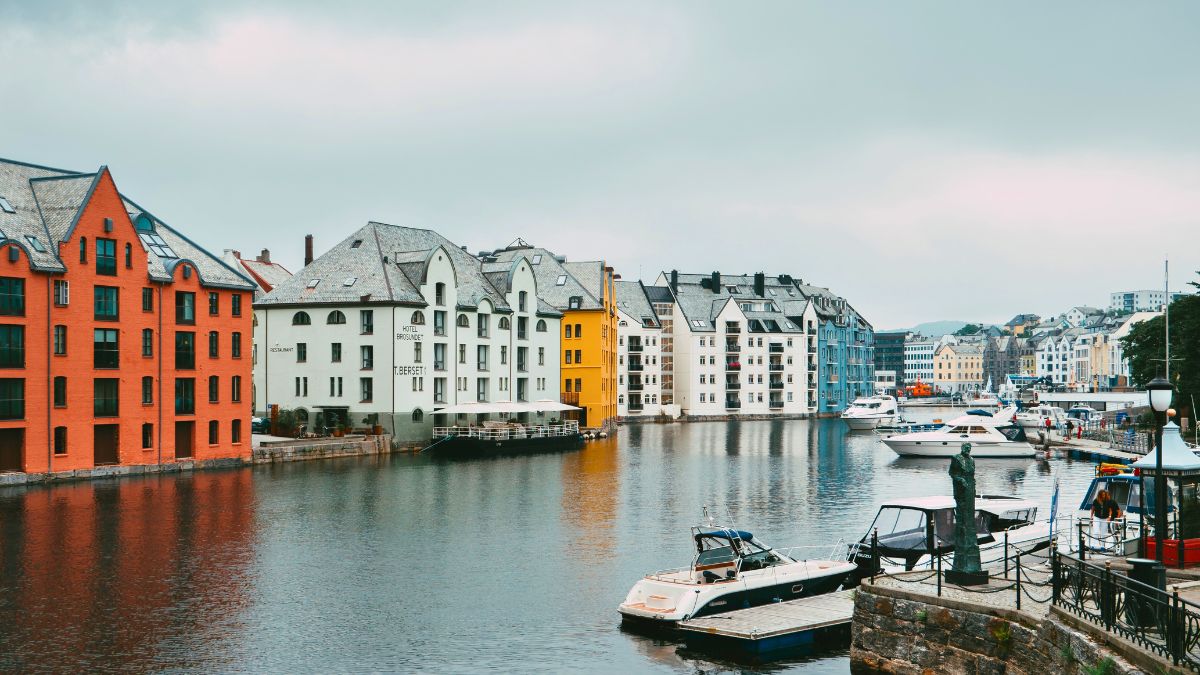 Colorful buildings line a serene waterfront, reflecting in the calm water. Several yachts are moored, while a cloudy sky looms overhead.
