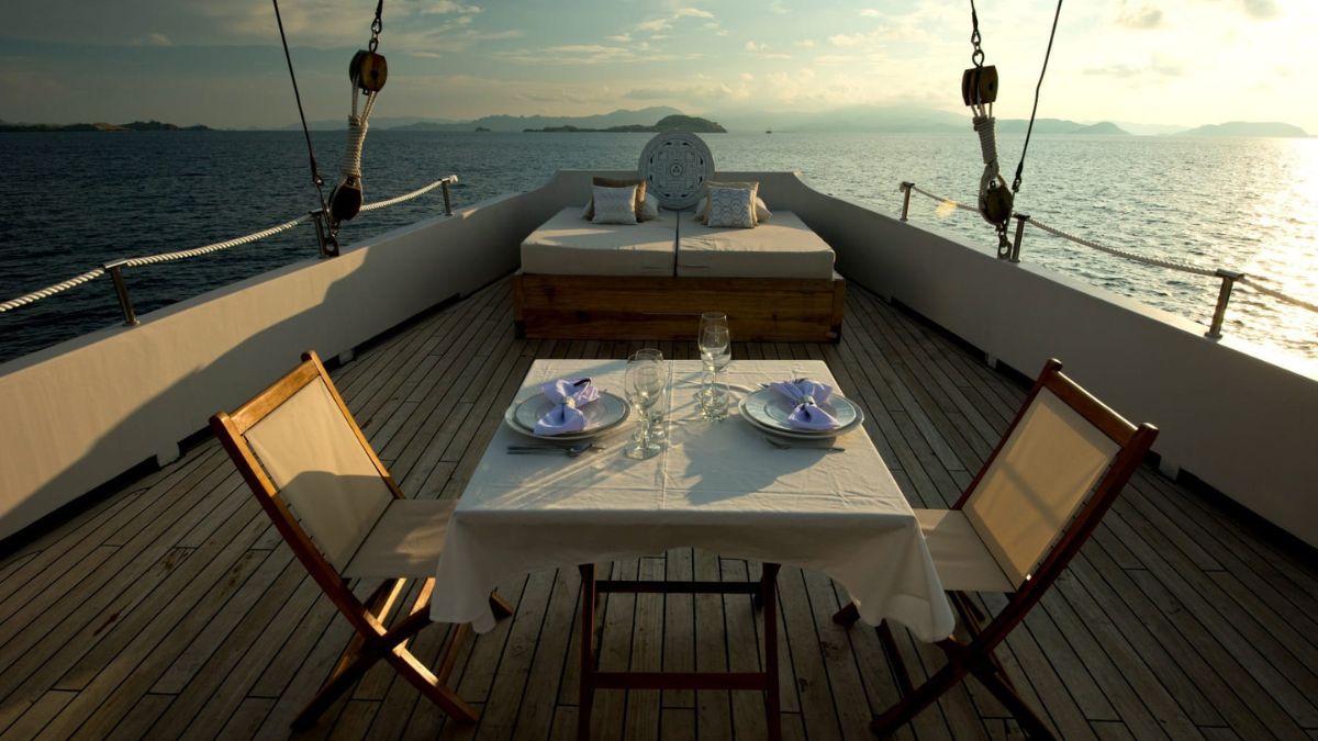 A beautifully set dining table with purple napkins overlooks calm water and distant islands. A lounge area with cushions is visible in the background, creating a serene atmosphere.