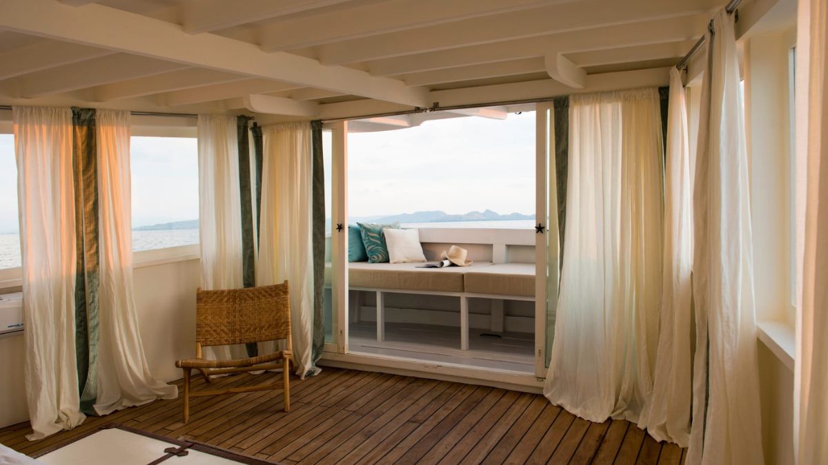 A cozy, light-filled room with wooden flooring and sheer white curtains. A comfortable daybed with pillows is visible through an open sliding door, offering a view of the water and distant hills. A natural wood chair is positioned nearby.