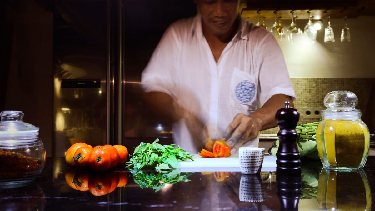 A person in a casual white shirt chops vegetables on a countertop. Fresh tomatoes, herbs, and spices are arranged nearby, with a glass jar of mustard and a pepper grinder in view. The kitchen background shows soft lighting and hanging glassware.