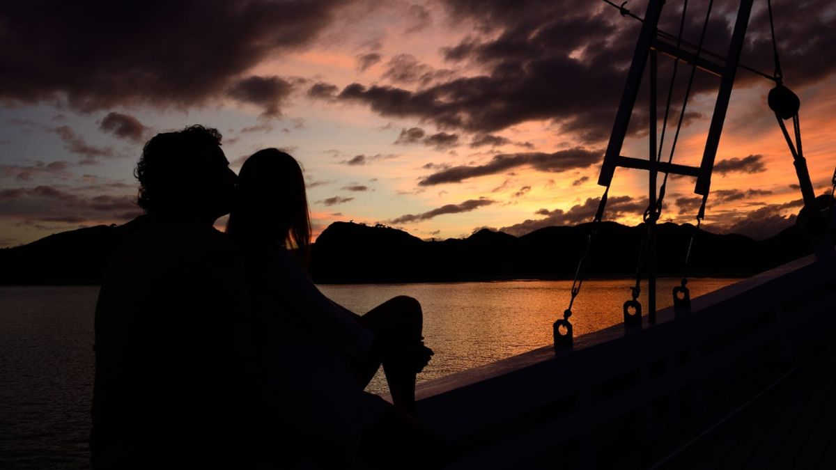 A couple sharing a kiss silhouetted against a vibrant sunset over calm waters, with mountains in the background and clouds creating a dramatic sky.