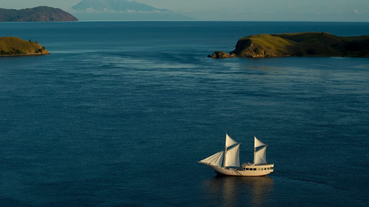 A majestic sailing ship with white sails glides across the calm blue waters, surrounded by distant island hills and a hazy mountain in the background.