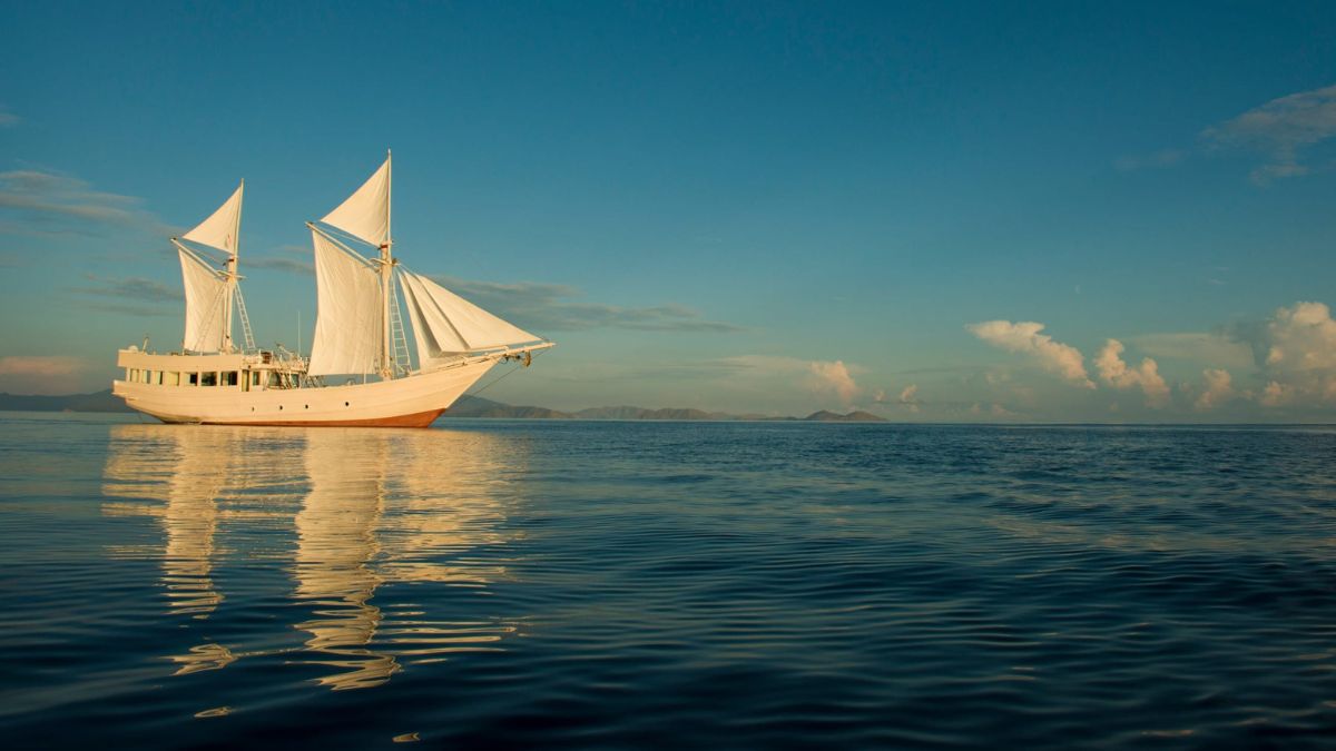 A large sailing ship with white sails glides across calm blue waters, reflecting its image. The sky is clear with a few fluffy clouds, and distant hills can be seen on the horizon.