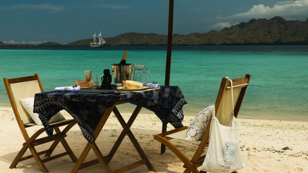 A beautifully arranged beachside dining setup with a table draped in a patterned cloth, featuring a bottle of champagne, glasses, and a plate of food. Two wooden chairs flank the table, set against a stunning backdrop of turquoise water and distant islands under a partly cloudy sky. A sailboat glides in the background.