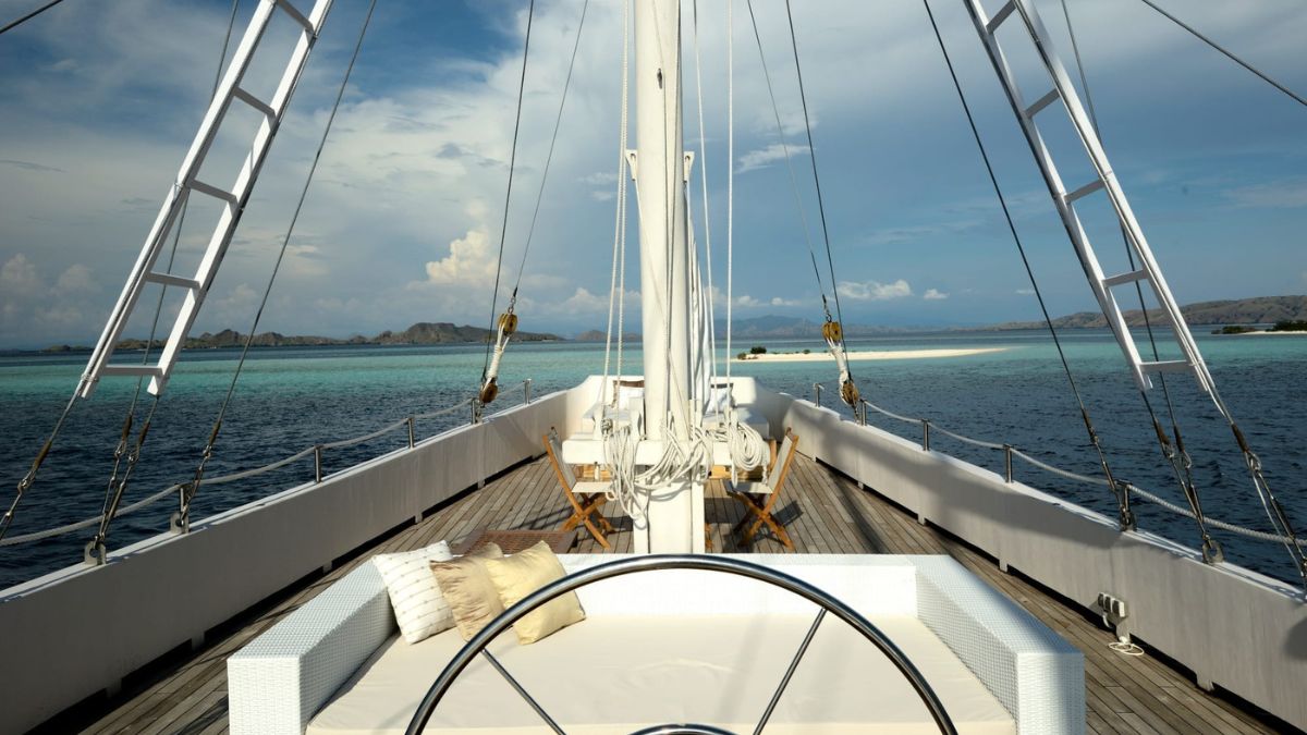 A spacious yacht deck with a large steering wheel in the foreground, surrounded by a white seating area. In the background, calm turquoise waters stretch towards distant islands under a partly cloudy sky.