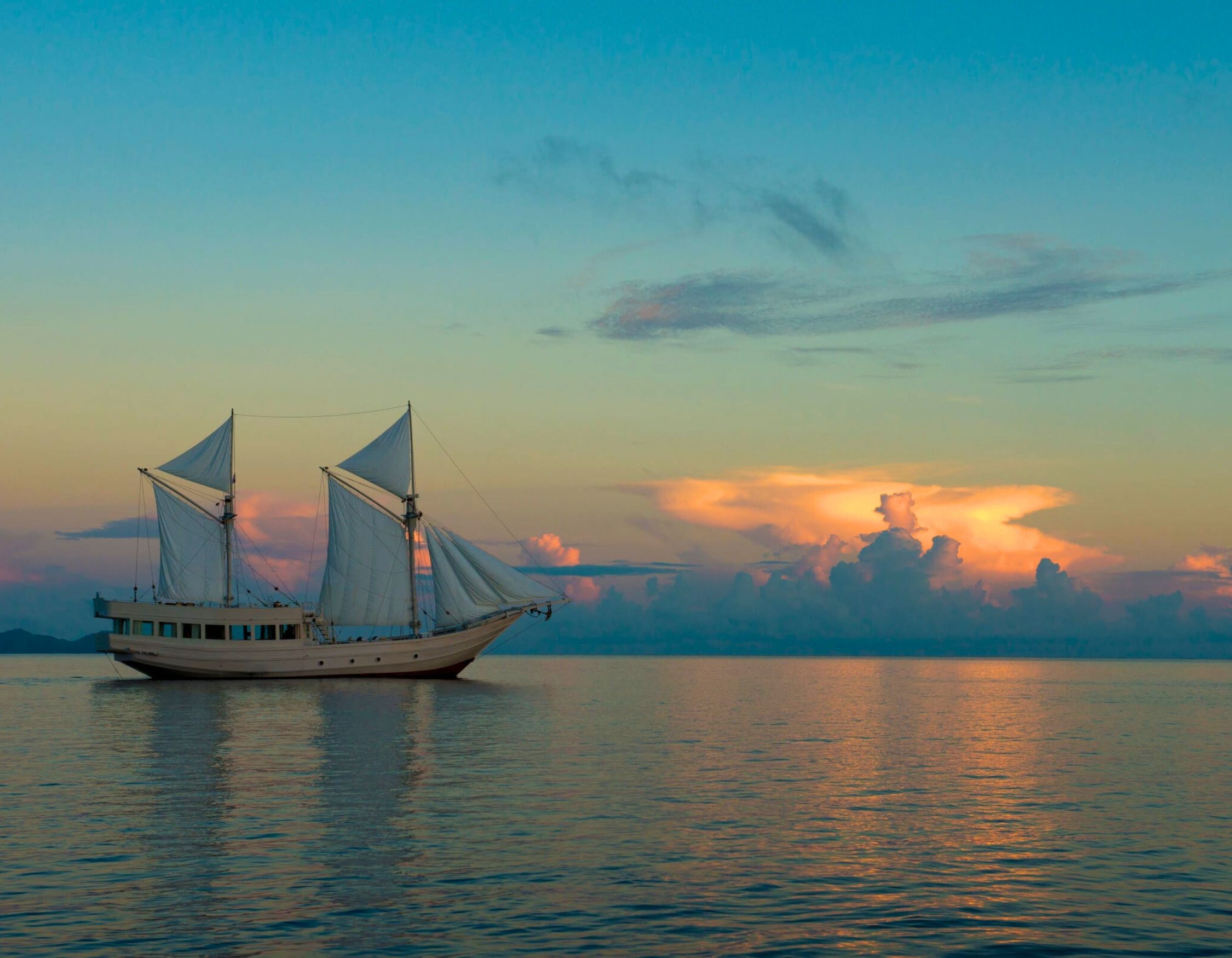 A traditional sailing ship with white sails glides on calm waters during sunset. The sky is a blend of warm hues with soft clouds, reflecting a serene atmosphere.