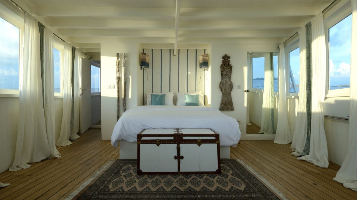A cozy, bright bedroom featuring a large bed with white bedding, decorative pillows, and a vintage trunk at the foot. Windows provide natural light and a view of the ocean, with wooden floorboards and elegant curtains completing the serene atmosphere.