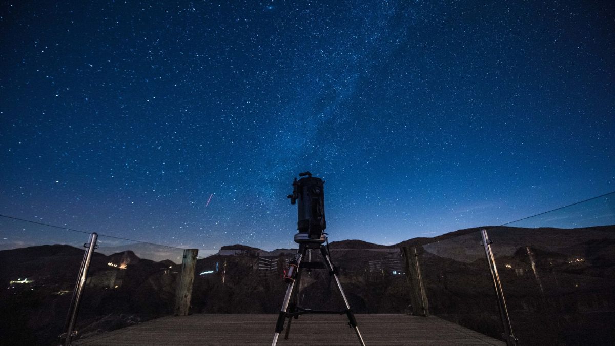 A telescope set up on a wooden platform under a starry night sky, with the Milky Way visible above and distant hills in the background.