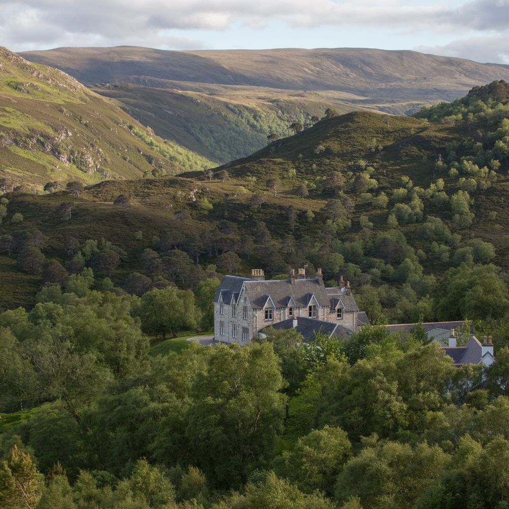 A large, elegant stone house surrounded by lush green trees and rolling hills. The mountainous landscape features a mix of greenery and rocky outcrops under a cloudy sky.