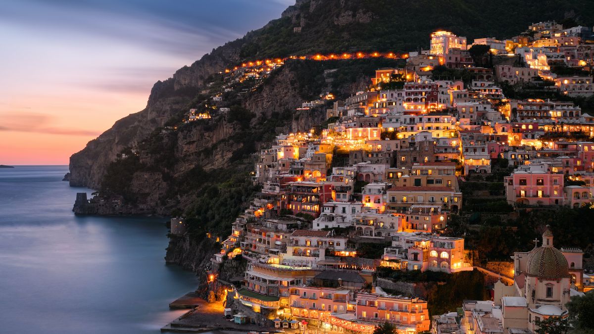 A scenic view of Positano at dusk, with brightly lit buildings cascading down a steep cliff towards the sea. The sky features soft hues of purple and orange as the sun sets on the horizon, reflecting gently on the water.
