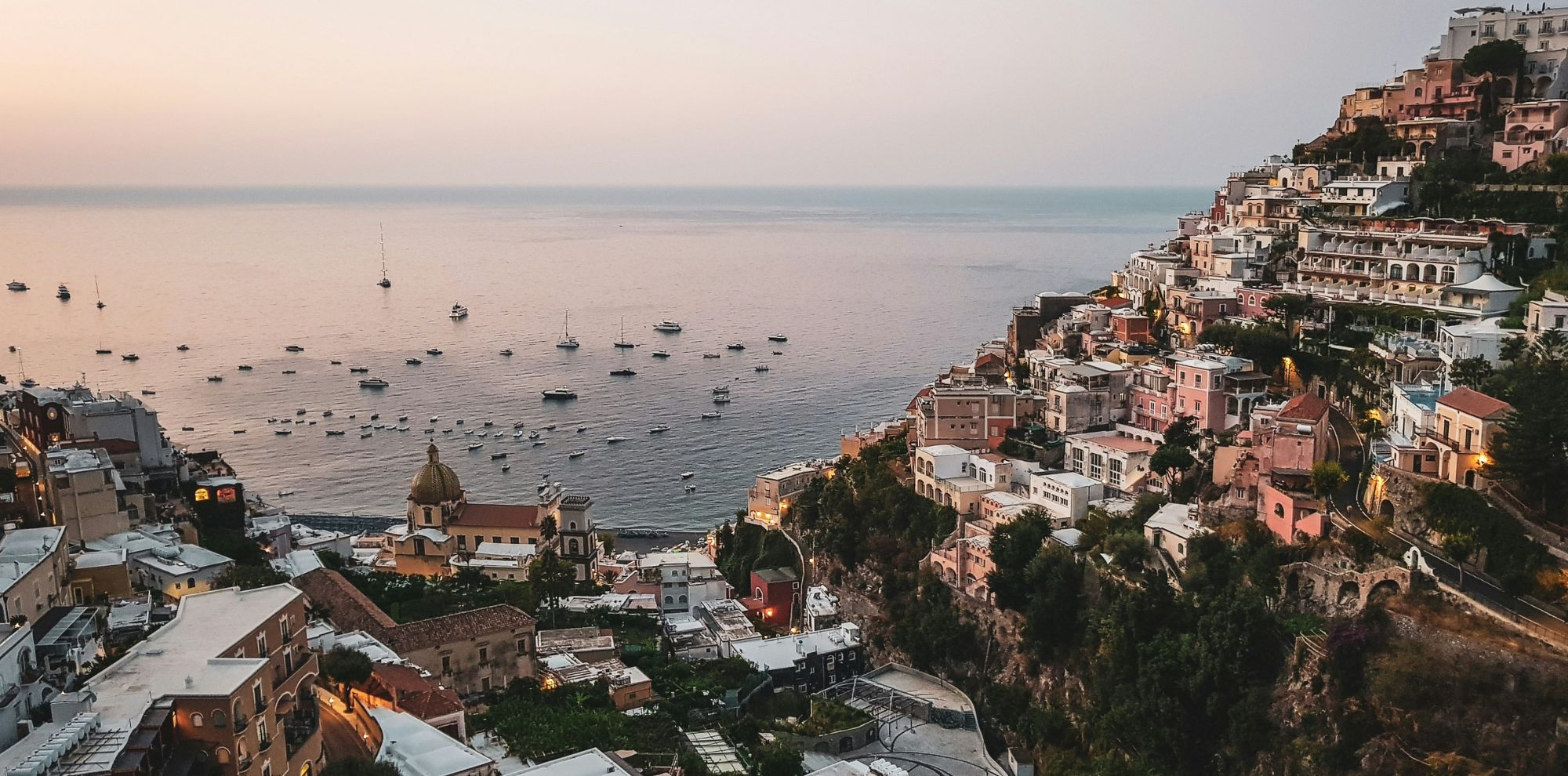 A scenic view of Positano, Italy, at sunset, showcasing colorful buildings perched on steep cliffs overlooking the calm sea filled with boats. The sky transitions from orange to blue as evening approaches.