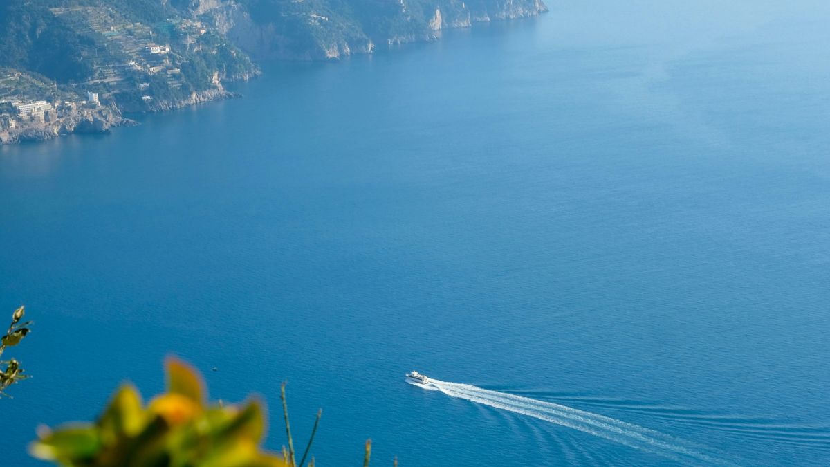 A small boat leaves a trail on the calm blue waters, set against steep coastal cliffs and a clear sky.