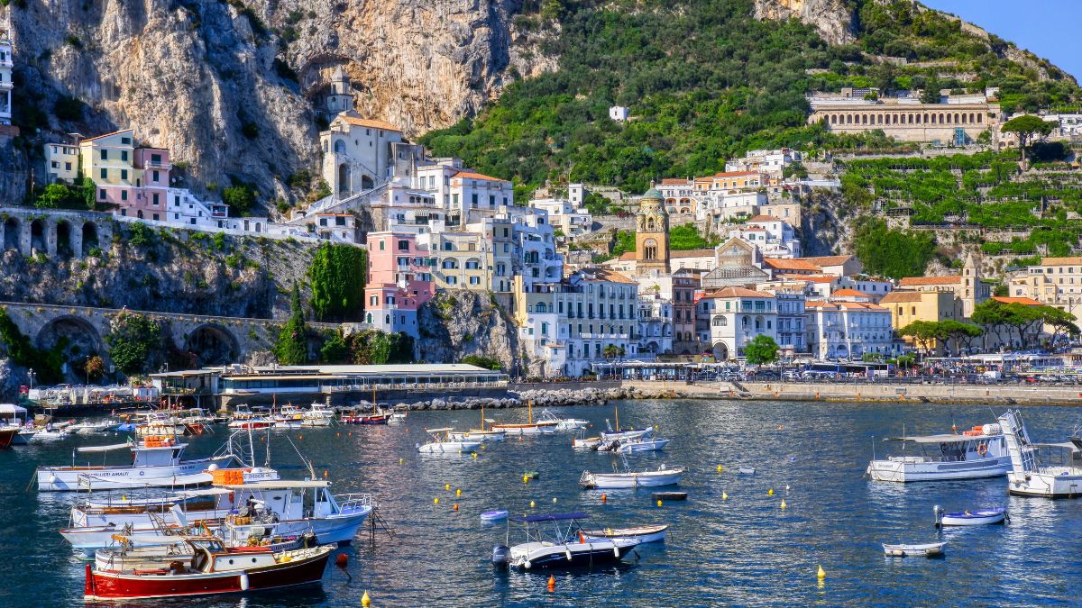 Colorful buildings line the cliffside above a calm bay, dotted with various boats. Lush greenery and rocky hills in the background provide a picturesque coastal scene.