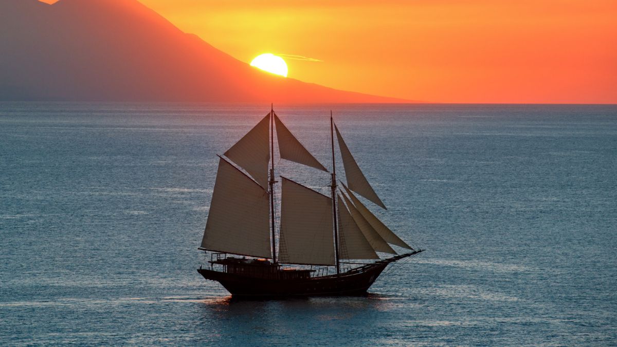 A sailing ship with tall masts glides across calm waters during a vibrant sunset, with the sun dipping behind a distant mountain.