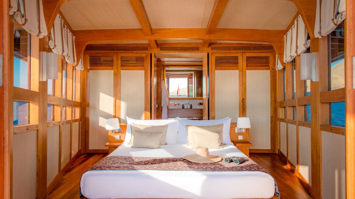 A cozy bedroom on a boat, featuring wooden walls and ceiling, large windows for natural light, a double bed with decorative pillows, a straw hat, and sunglasses on the bed. Nautical decor is present with a glimpse of the ocean visible through the windows.