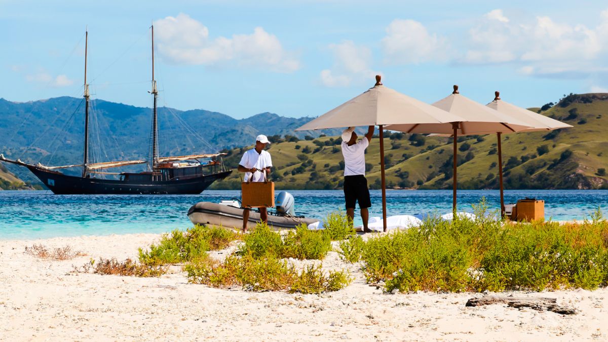 A sandy beach with two men setting up umbrellas and preparing a small area near a boat. In the background, a large wooden ship is anchored in turquoise waters, with lush green hills and a blue sky.