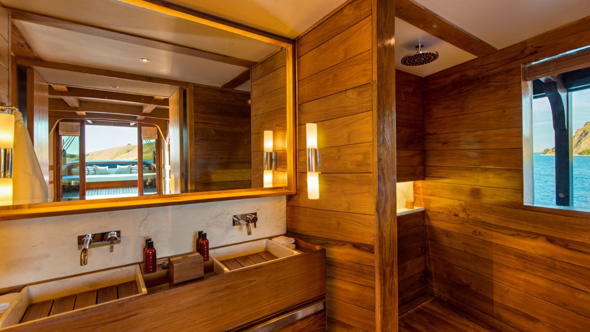 A luxurious wooden bathroom featuring dual sinks with sleek faucets, a large mirror, and natural light streaming through a window. The warm wood paneling creates a serene ambiance, complemented by elegant lighting fixtures.