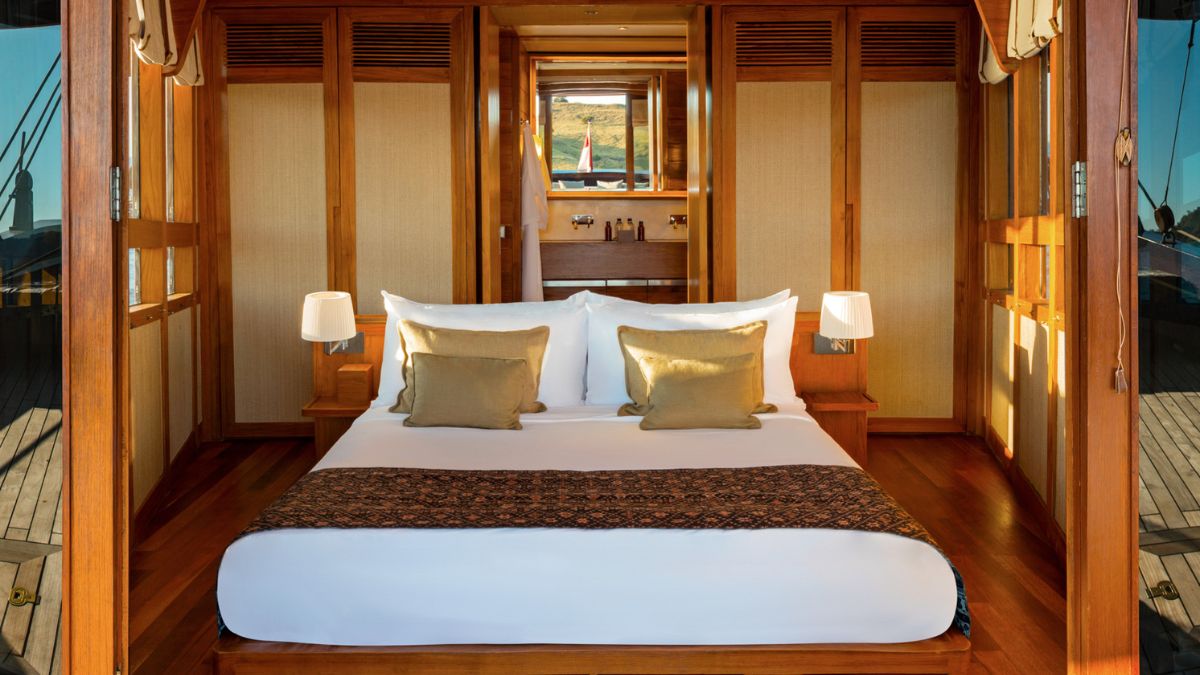 A luxurious wooden cabin interior featuring a large bed with white linens and decorative pillows. Soft lighting from bedside lamps creates a warm atmosphere. The room is adorned with elegant wood paneling and has large windows allowing natural light to fill the space.