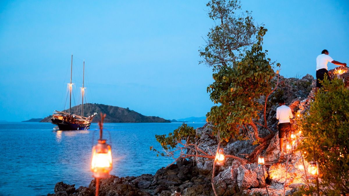 A ship anchored near a rocky shore at dusk, with lanterns illuminating the surrounding area and a person climbing up the rocks. Soft blue hues fill the sky, and a distant island is visible on the horizon.