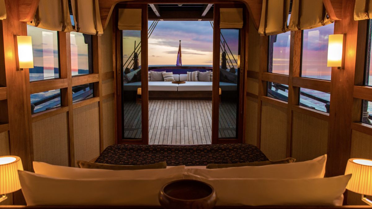 A cozy cabin interior featuring wooden walls and large windows with a view of a sunset over the ocean. The room includes a plush bed with decorative pillows and soft lighting, leading to a spacious outdoor lounge area.