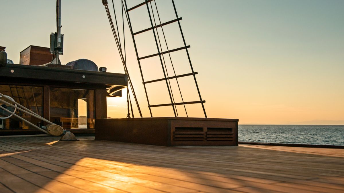 A wooden deck of a ship captures the golden glow of sunset, with a metal ladder leading upward against the backdrop of the calm sea.