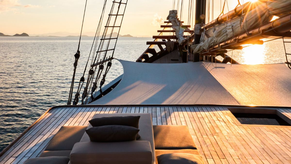 A luxurious yacht deck at sunset, featuring sleek wooden flooring, sun loungers, and the silhouette of sails and rigging against a tranquil ocean backdrop.