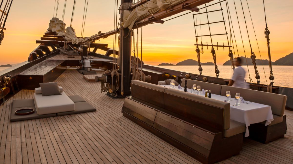 A beautifully designed dining area on a sailing yacht, featuring a long table set for a meal with glasses and a tablecloth. The deck is made of polished wood, and a lounge area with cushions is visible. The backdrop showcases a stunning sunset over the water with silhouetted hills.