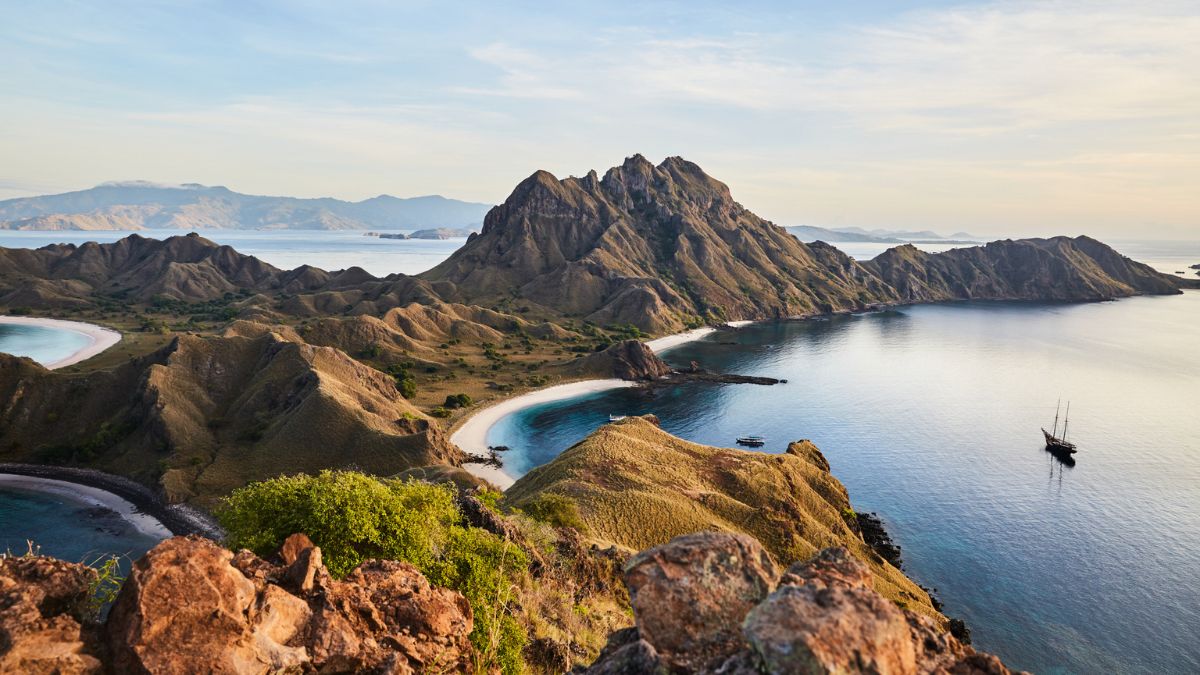 Breathtaking view of rugged mountains surrounding a calm bay, with white sandy beaches and a distant sailboat. The landscape features lush greenery and soft rolling hills, bathed in golden sunlight under a clear sky.