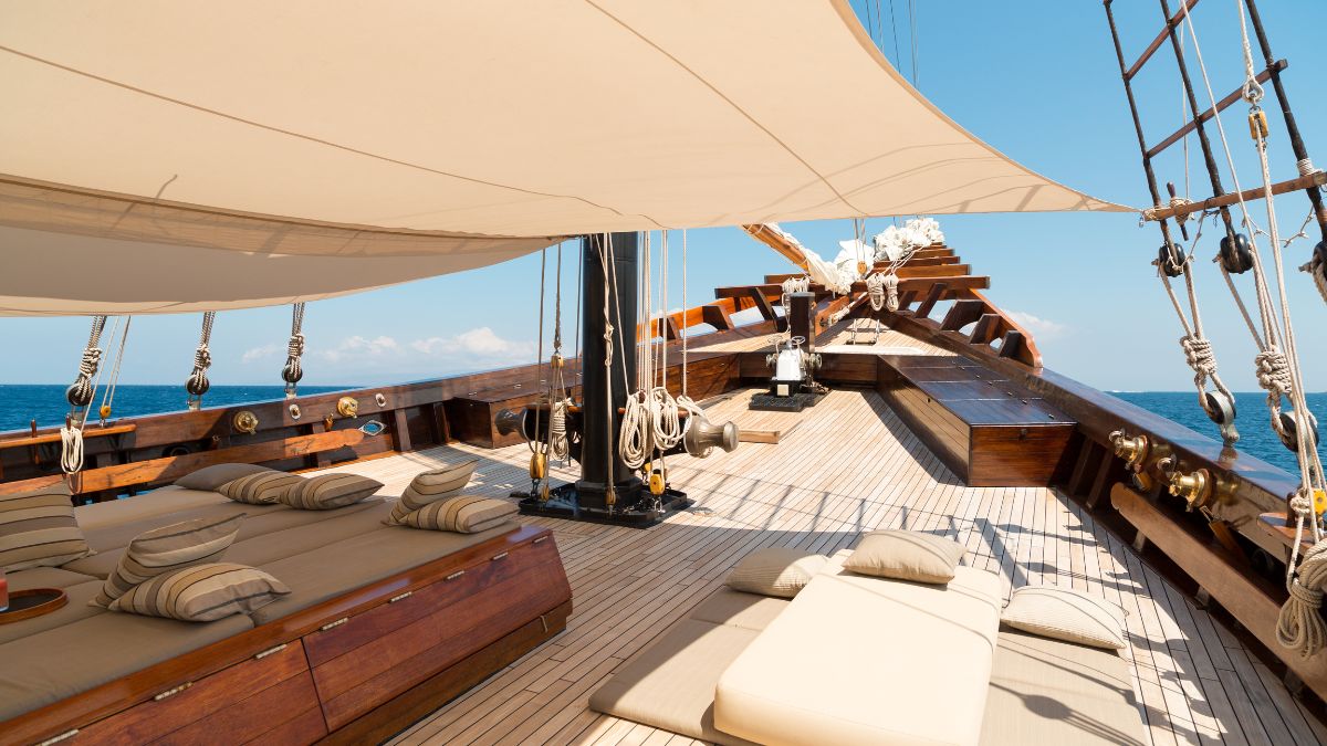 Spacious wooden deck of a sailing yacht, featuring plush sunbeds and cushions, with sails unfurled against a clear blue sky and calm sea.
