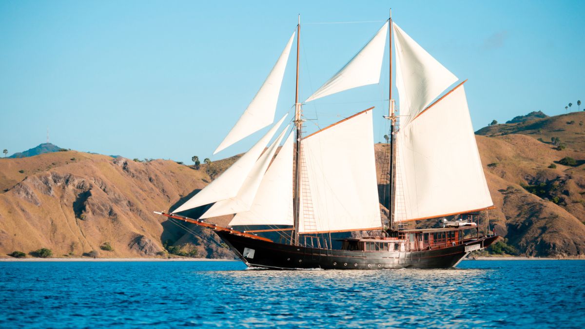 A tall ship with white sails glides across blue water, set against a backdrop of rugged, green hills.