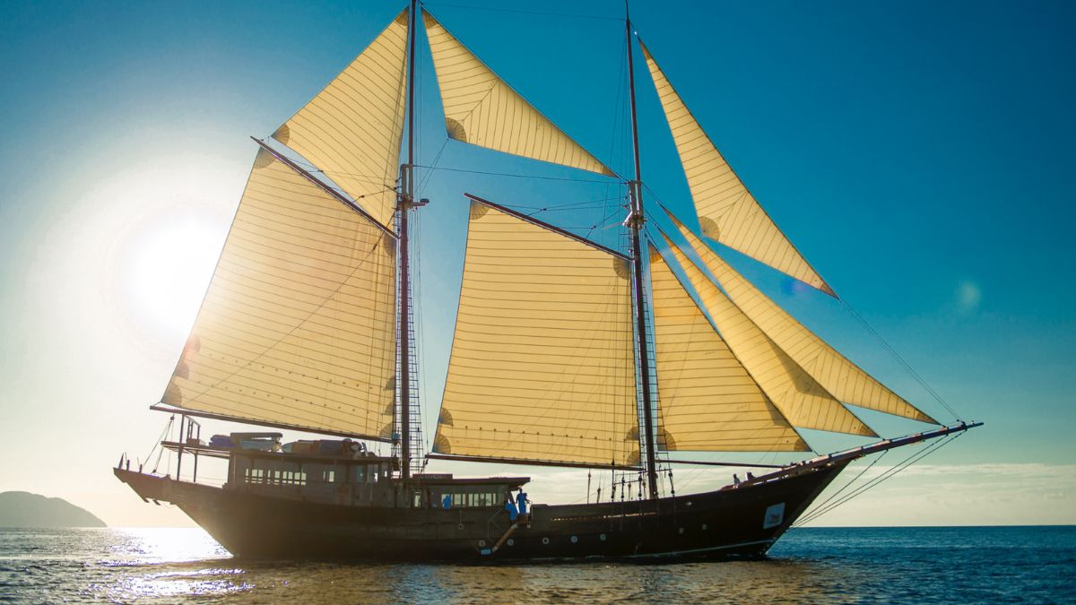 A large sailboat with prominent yellow sails gliding over calm waters, set against a bright blue sky and a glowing sun in the background.