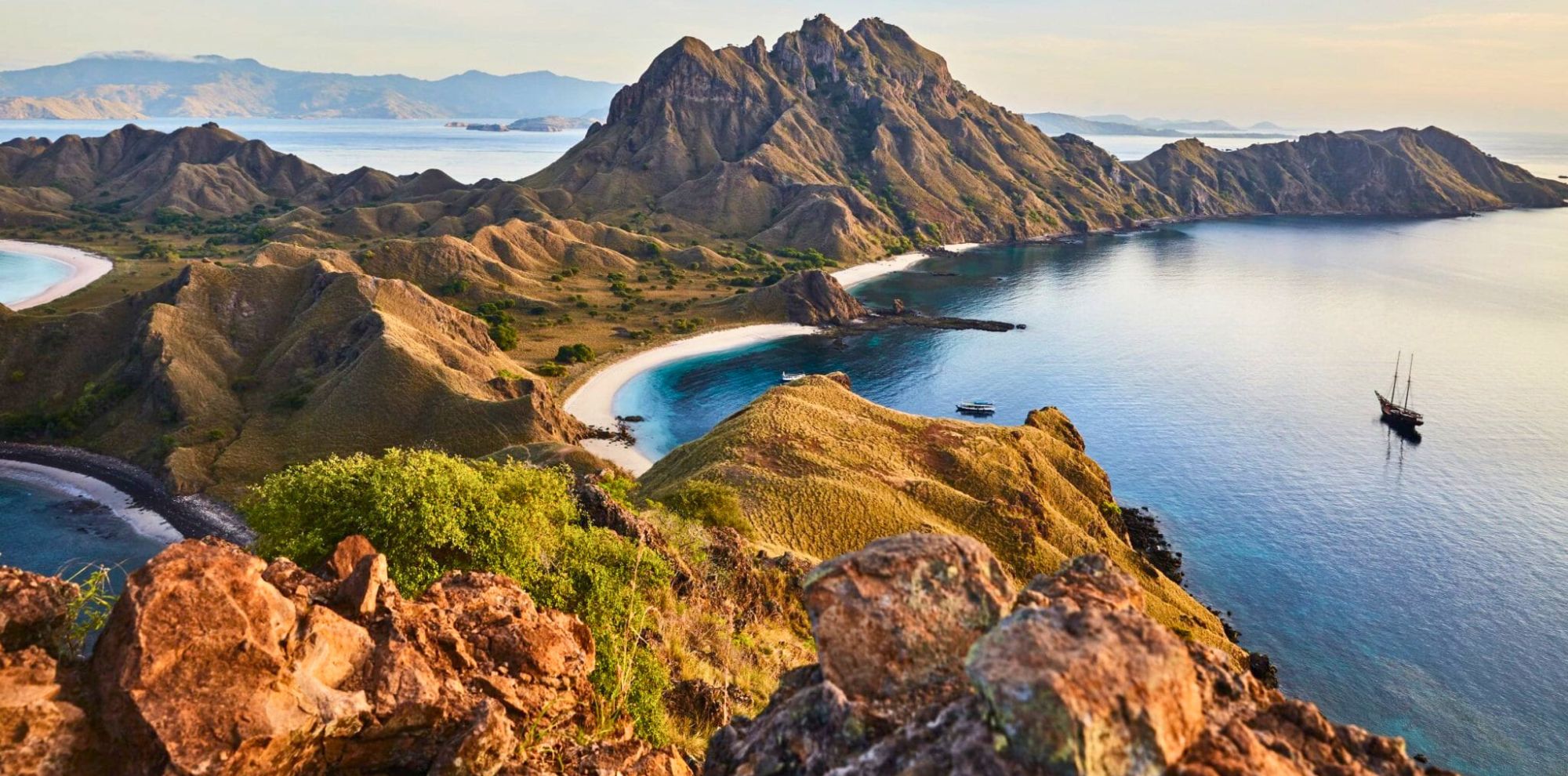 A stunning view of a rugged coastal landscape featuring rolling hills, a sandy beach, and calm blue waters. In the distance, several islands are visible under a clear sky, while a tall ship rests in the water near the shore.