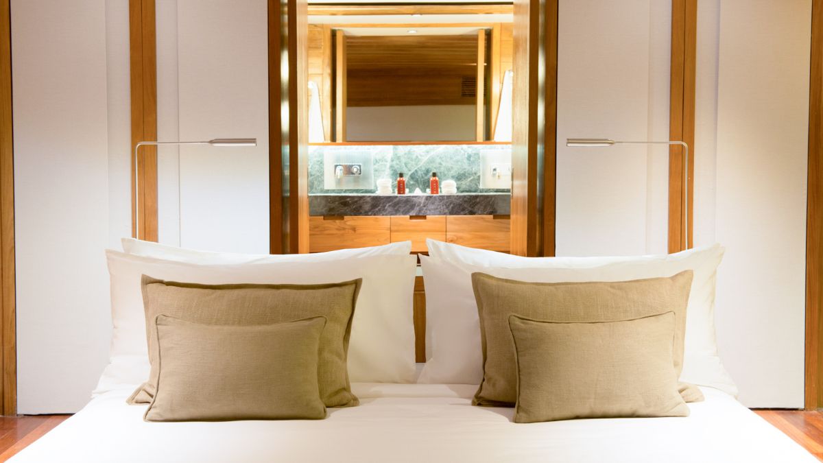 A modern, well-lit bedroom featuring a neatly made bed with two large white pillows and decorative beige pillows. In the background, a stylish bathroom with marble accents is visible through a mirrored view.