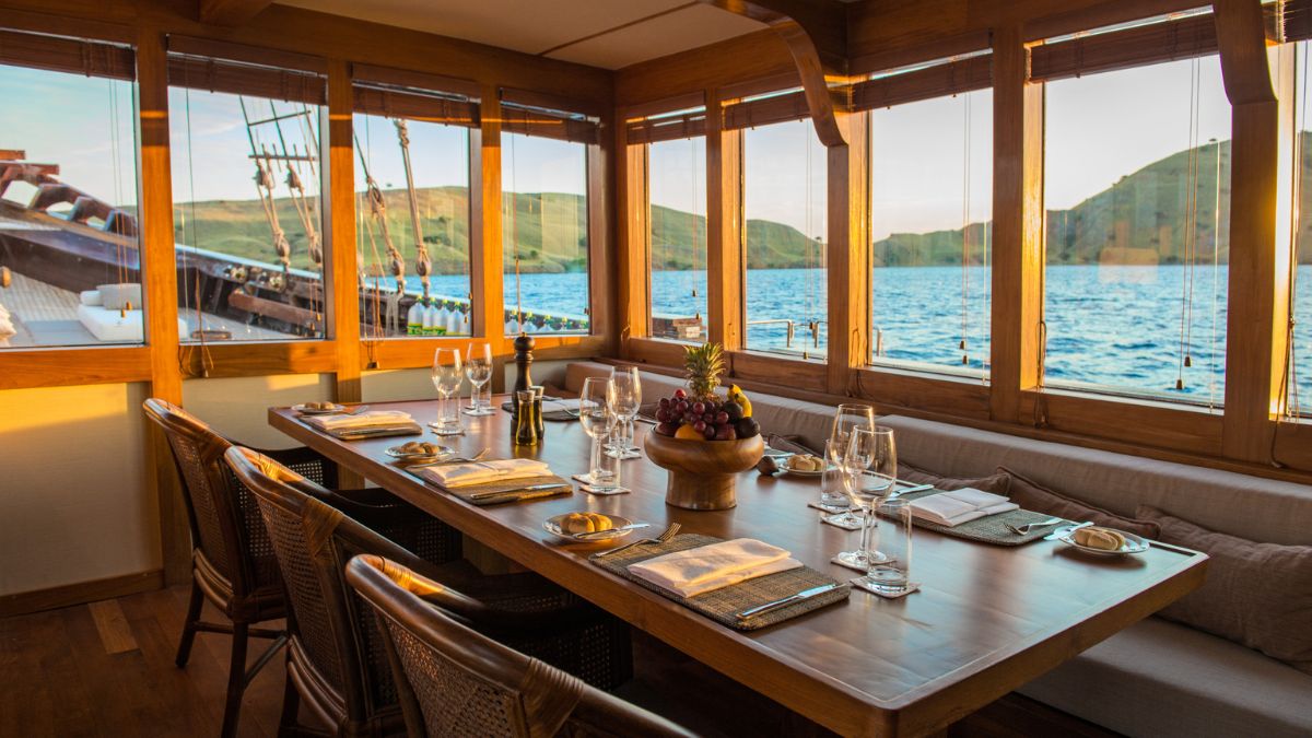 A beautifully set dining table with glasses, cutlery, and plates, surrounded by wooden chairs. The large windows offer a scenic view of rolling hills and the sea, with warm sunlight streaming in. A fruit bowl centerpiece adds a touch of elegance.