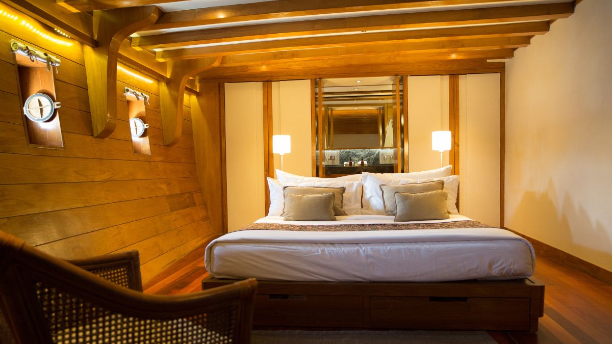 A cozy bedroom featuring a large bed with white linens and decorative pillows. Wooden walls and beams create a warm atmosphere, complemented by soft lighting from wall sconces. A chair is positioned near the bed, adding to the inviting decor.