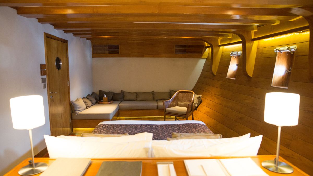 A cozy yacht cabin featuring wooden walls and ceiling, with a queen-sized bed, comfortable seating area, and soft lighting from lamps. There are porthole windows and tasteful decor, creating a warm and inviting atmosphere.