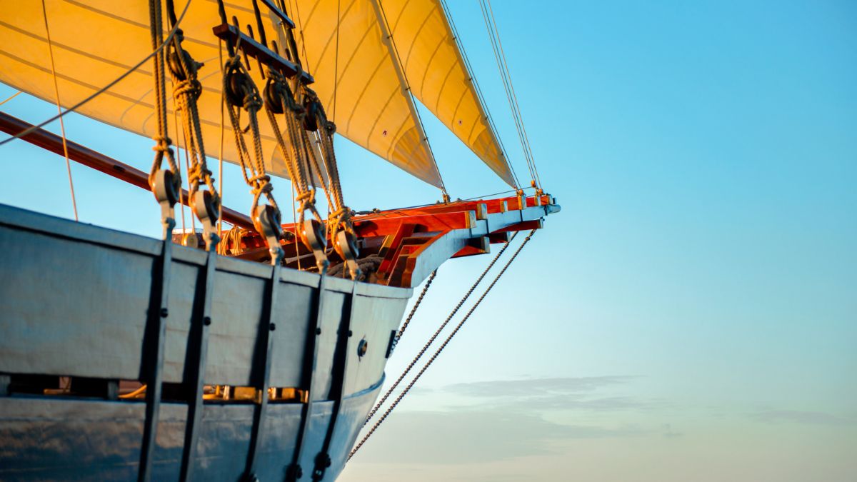 A sailing ship with bright yellow sails against a clear blue sky. The ship's bow is prominently displayed, showcasing its wooden structure and rigging.