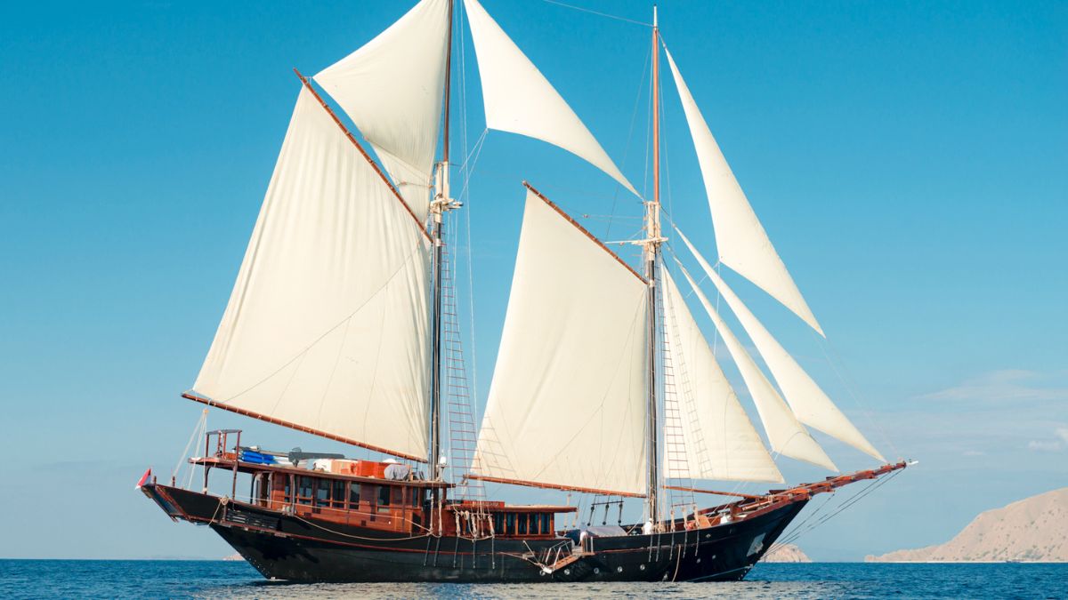 A large, traditional sailing ship with tall white sails gliding across calm blue waters, surrounded by a clear sky. The vessel features a dark wooden hull and various deck amenities.