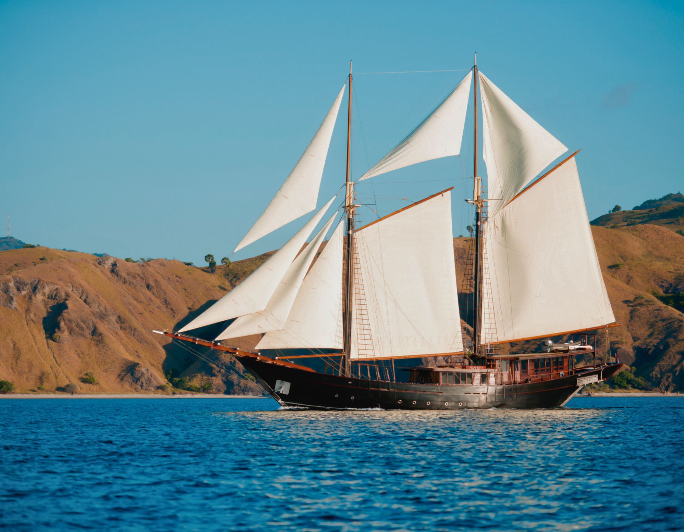 A majestic sailing ship with white sails glides through calm blue waters, accompanied by green hills in the background.