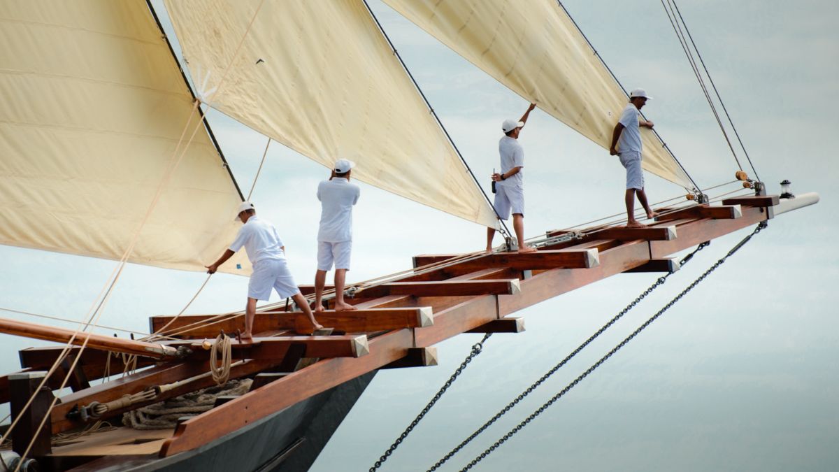 Sailors adjust the sails of a large boat, wearing white uniforms, as they work together on a wooden deck. The sky is overcast, creating a calm atmosphere.