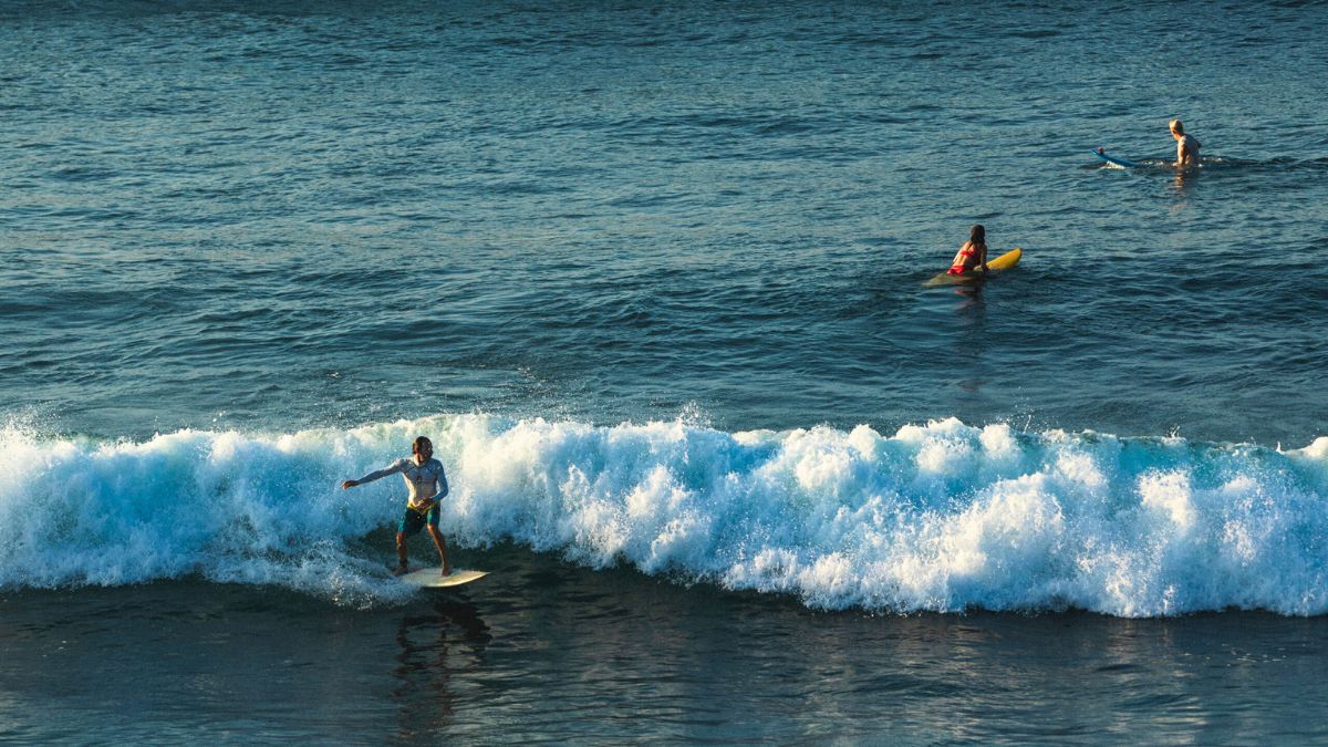 A surfer riding a wave in the foreground, while two people on surfboards are further back in the water. The ocean is a vibrant blue with frothy white waves.