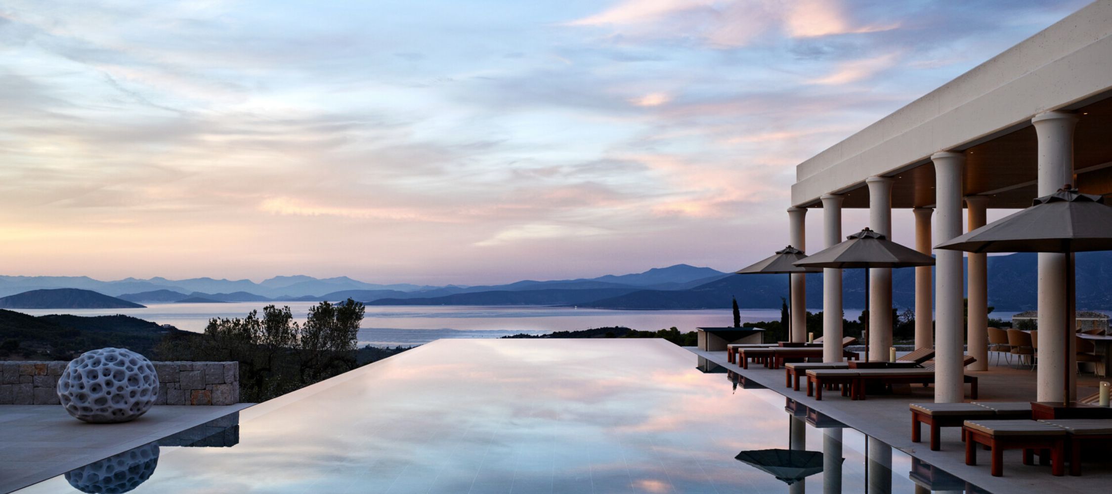 A modern villa features an infinity pool that reflects the evening sky, surrounded by scenic mountains and calm waters. Lounge chairs and shaded umbrellas provide a relaxing outdoor space.