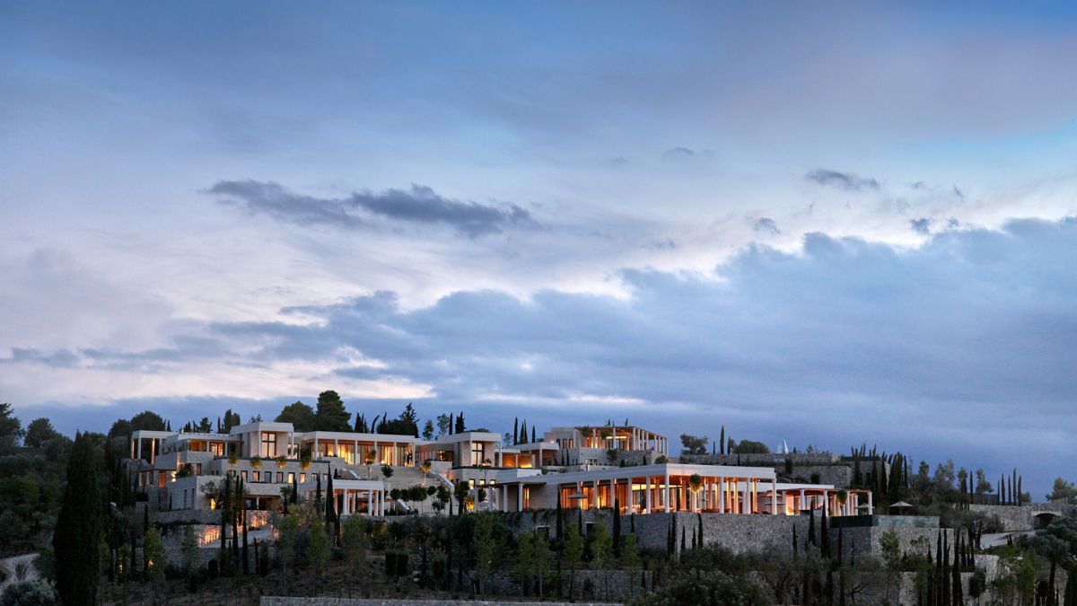 Modern hillside resort showcasing multiple elegant buildings with large windows amidst lush greenery and a cloudy sky at dusk.